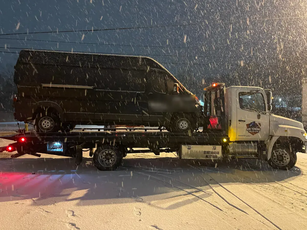 Joe Towing flatbed carrying a black van at night while snow falls.